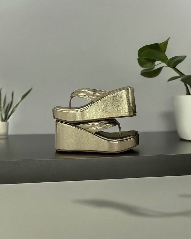 Pair of gold wedge platform slides with simple straps on a dark surface next to green potted plants
