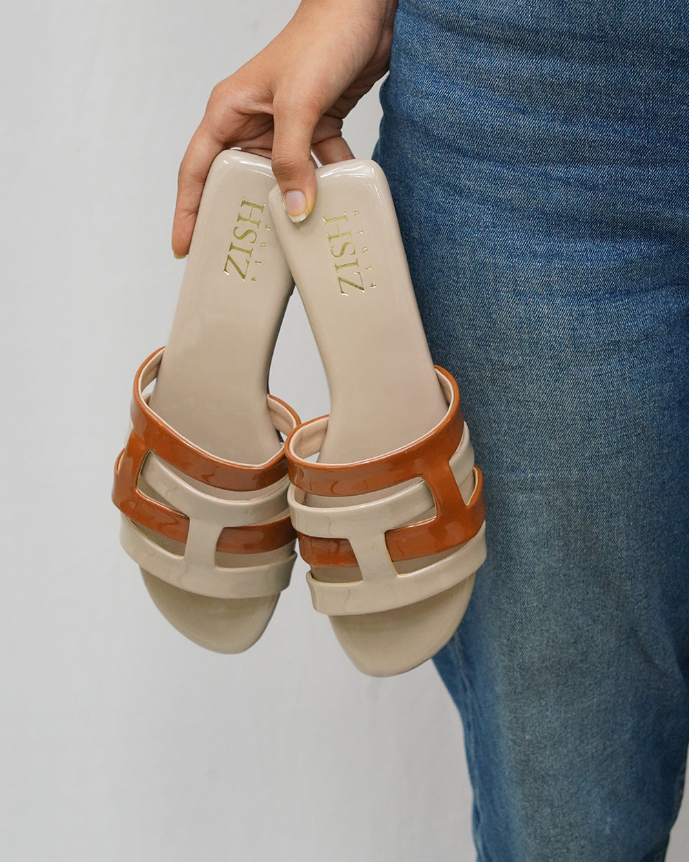 Beige Azzura Flats with tan geometric design open-toe sandals in PU vegan leather by Zish, held by hand against denim background