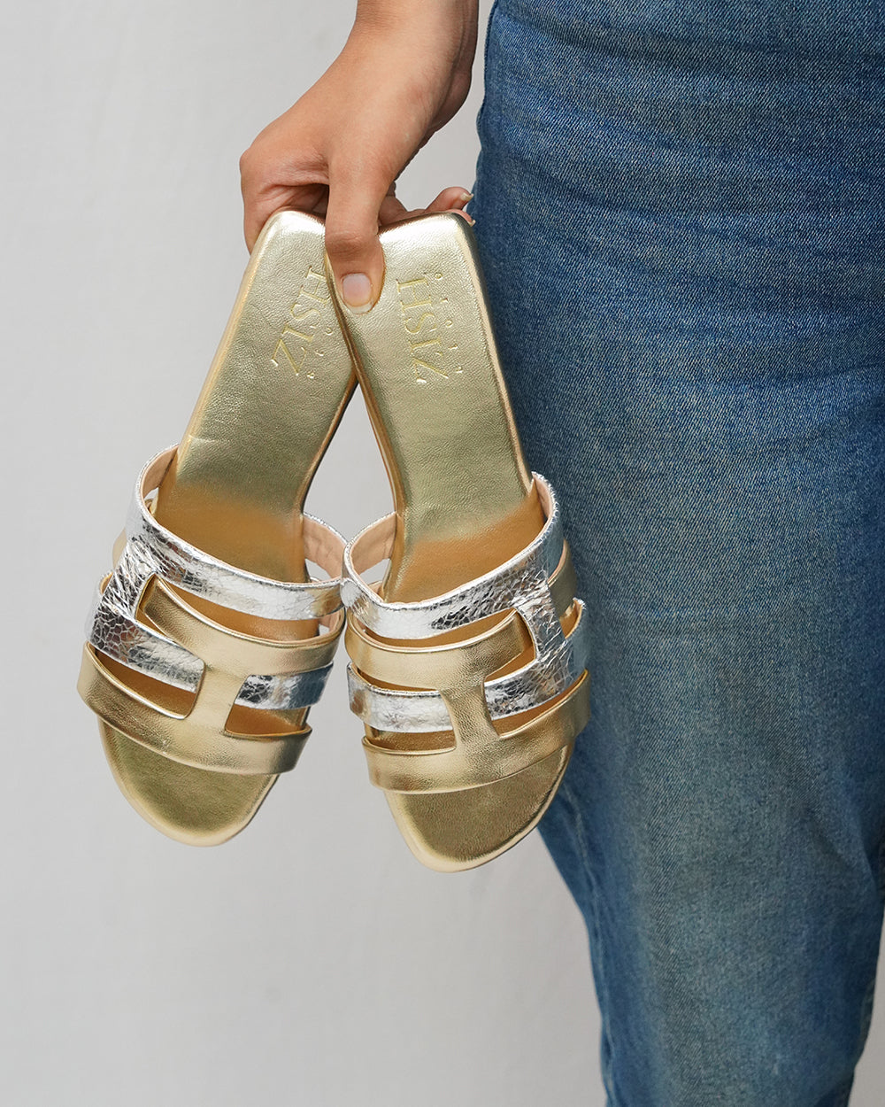 Hand holding Gold Azzura Flats featuring gold and silver geometric design, worn with blue denim skirt.