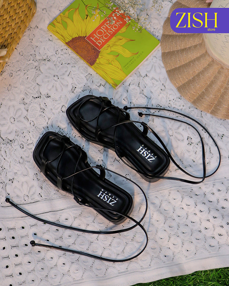 Black platform tie ups flats with thin straps on a white lace cloth background with a book and a straw hat nearby