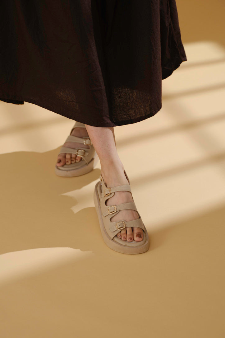 Beige triple buckle sandals with adjustable straps and cushioned footbed worn with a dark skirt on beige floor